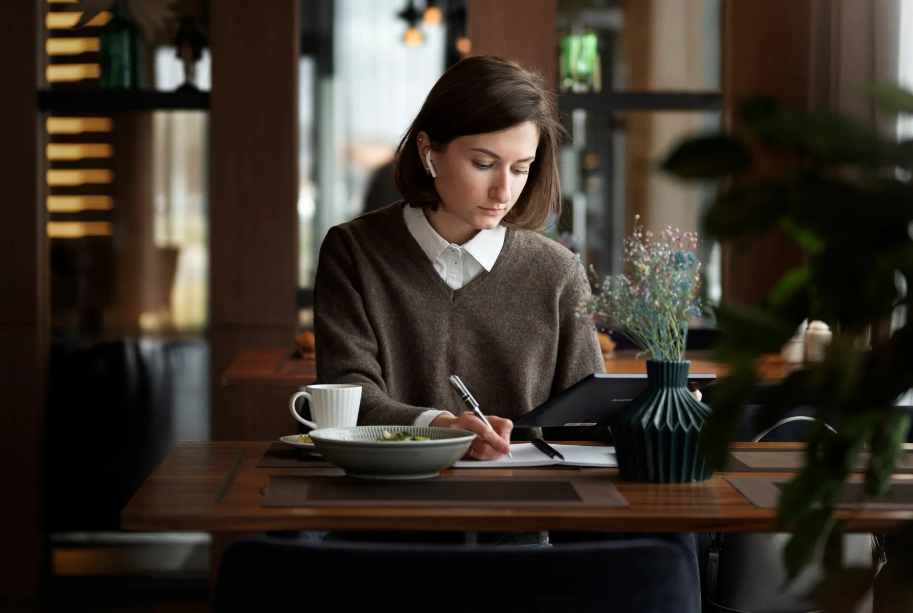 Young woman working in a cafe, writing in a notebook while using a tablet, with a coffee cup and meal on the table.